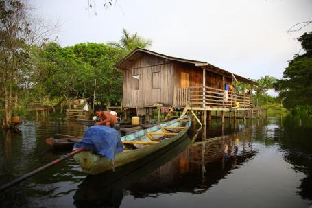A person navigates a large canoe next to a floating house in the Amazon rainforest.
